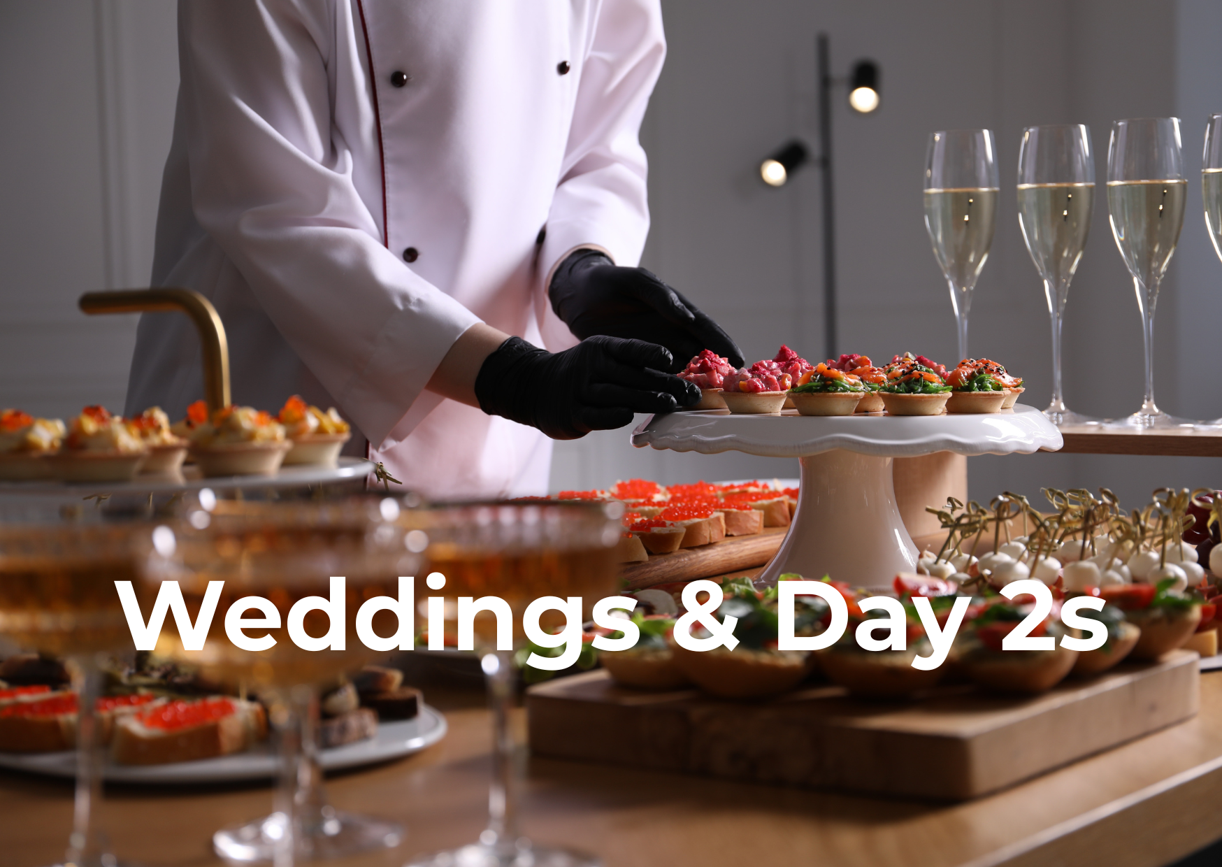 Chef in white uniform and black gloves arranging colorful appetizers on a table with champagne glasses, set for a wedding celebration.