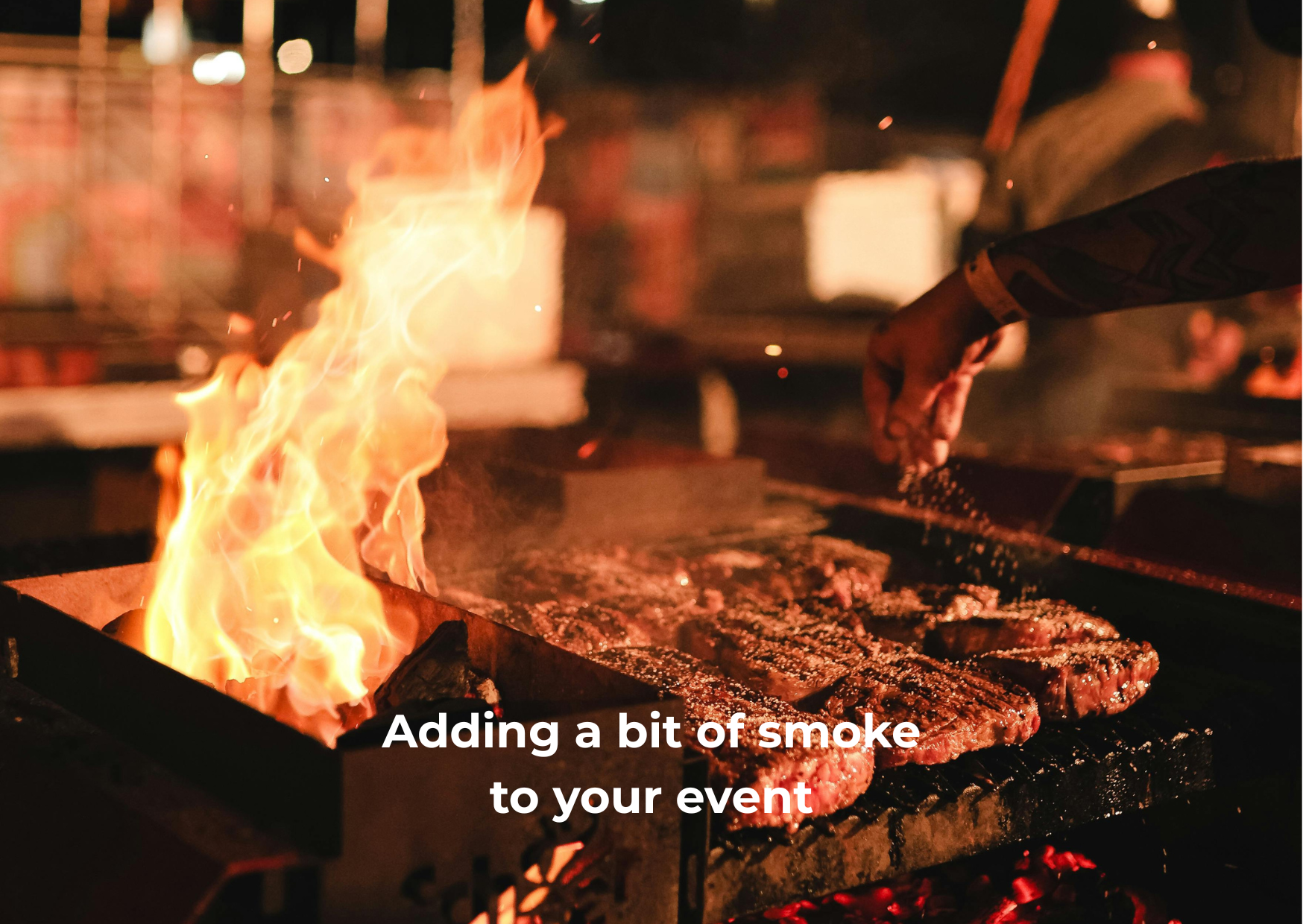 Close-up of steak being grilled over open flame, with flames and smoke rising, and a hand sprinkling seasoning on the meat, in a barbecue setting at night.