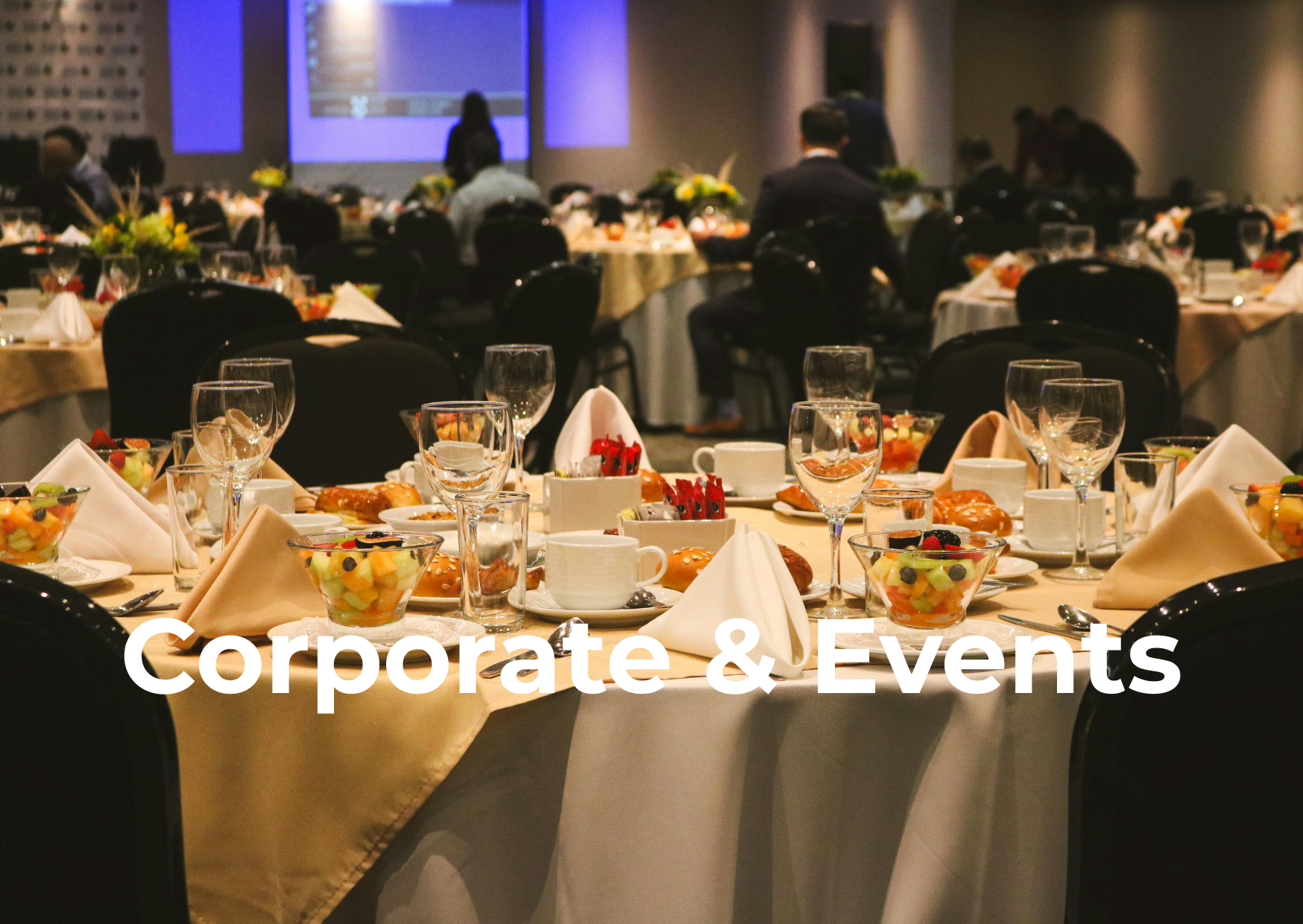 Banquet hall set up for a corporate event with round tables, china, glassware, and centerpieces, and a presentation screen in the background.