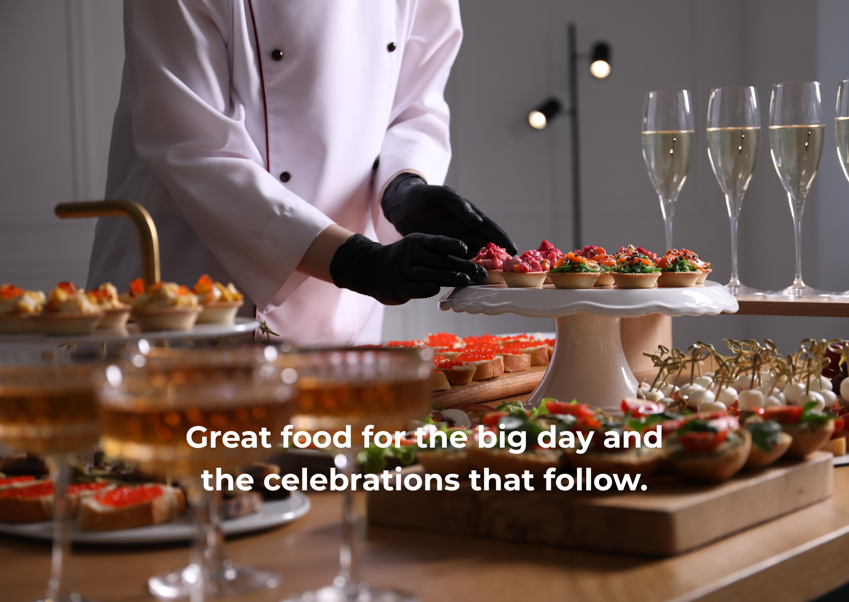 A chef in a white coat and black gloves preparing small appetizer bites on a dessert table with champagne glasses in the background.