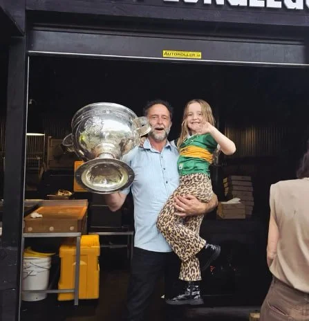 A man holding a young girl, both smiling, in front of a building with a sign that reads 'FREELING.' The man is holding a large trophy or glass bowl, and the girl is waving. The setting appears to be outdoors, possibly at an event or celebration.