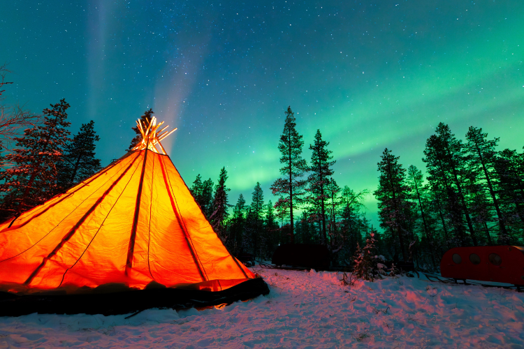 A lit tent beneath the northern lights in a snowy landscape.