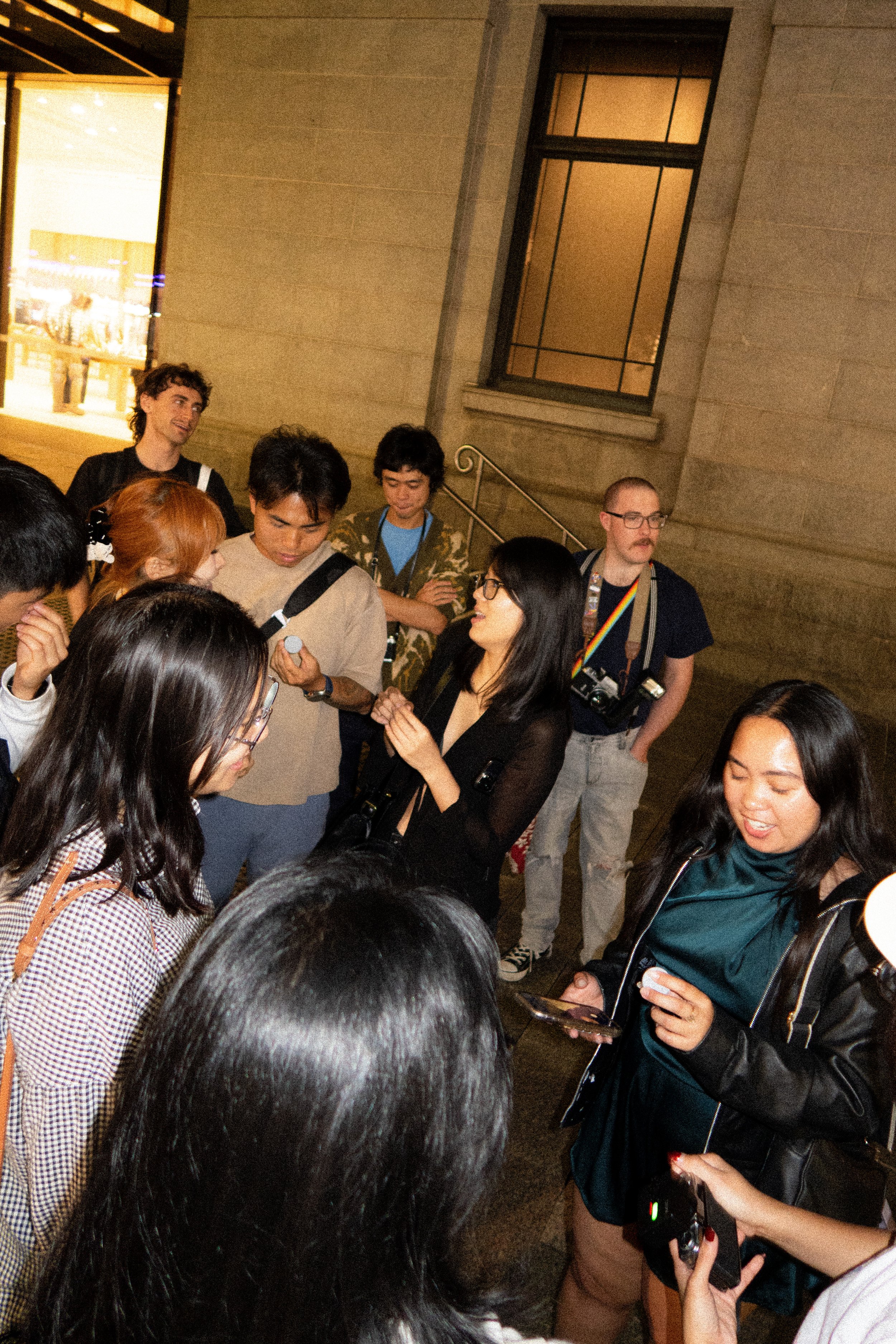 A group of young creatives gathered outdoors at night, chatting closely together while holding cameras and phones, capturing moments and connecting.