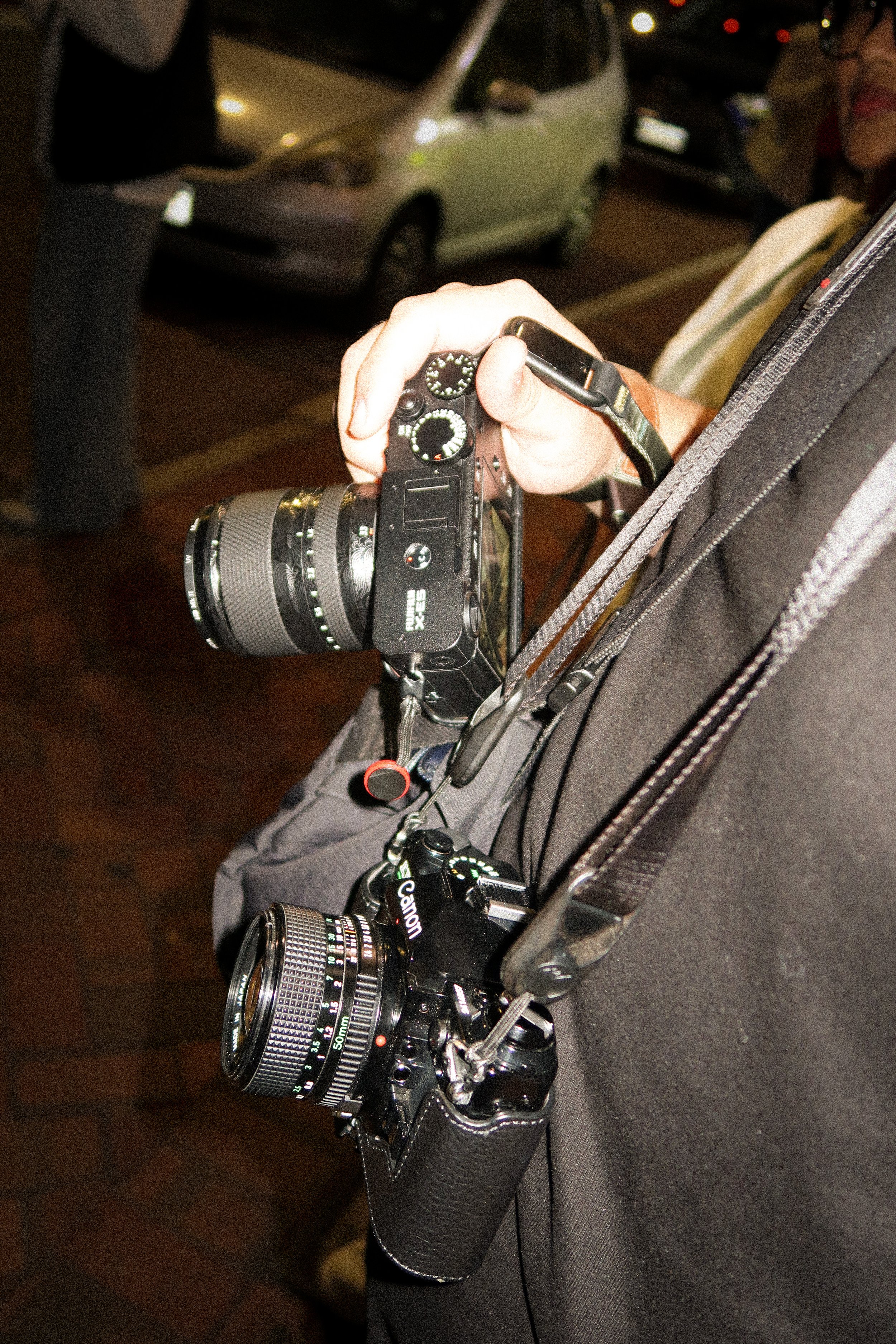 Close-up of a photographer holding a camera with another camera hanging from their shoulder at night.