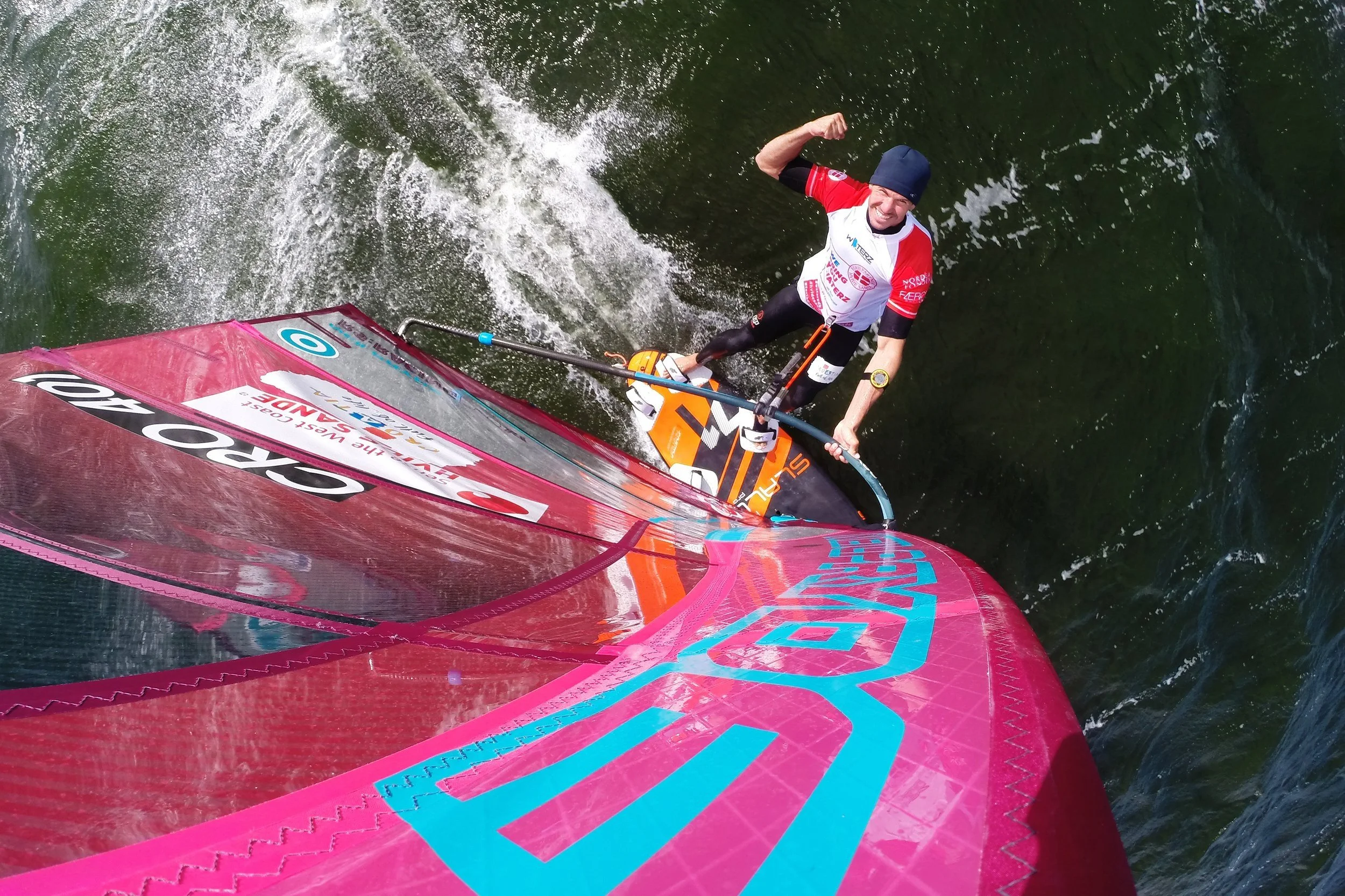 Person windsurfer på vand, set oppefra, iført rød og hvid dragt, stor pink sejl med tekst.