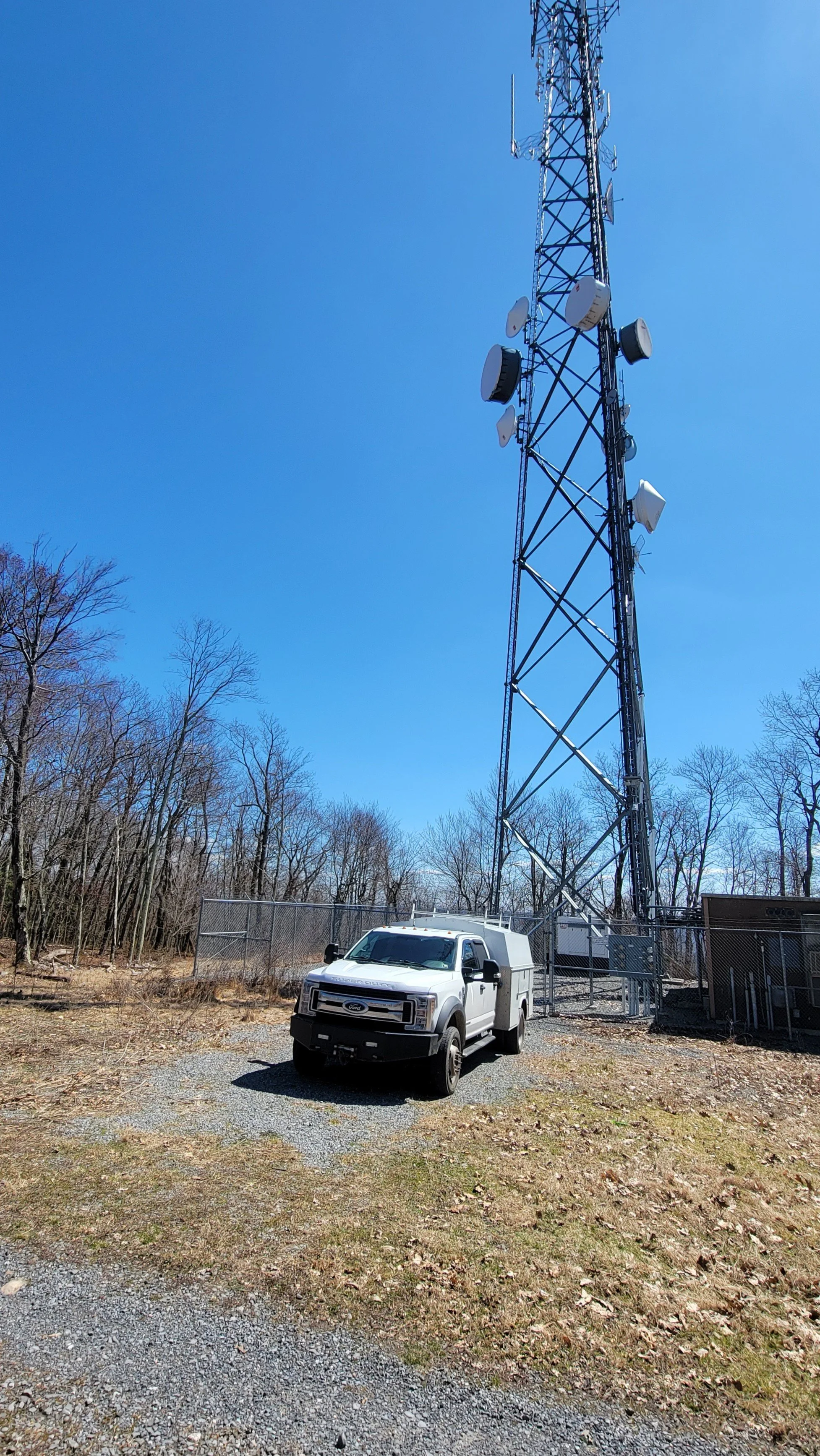 A tall radio tower with multiple antennas stands in a clearing surrounded by leafless trees. A white utility van is parked nearby on a gravel surface, enclosed by a chain-link fence.