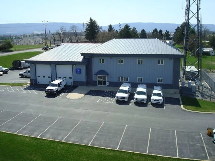 A gray industrial building with a metal roof, multiple garage doors, and a parking lot with several white trucks and cars, surrounded by greenery and power lines.