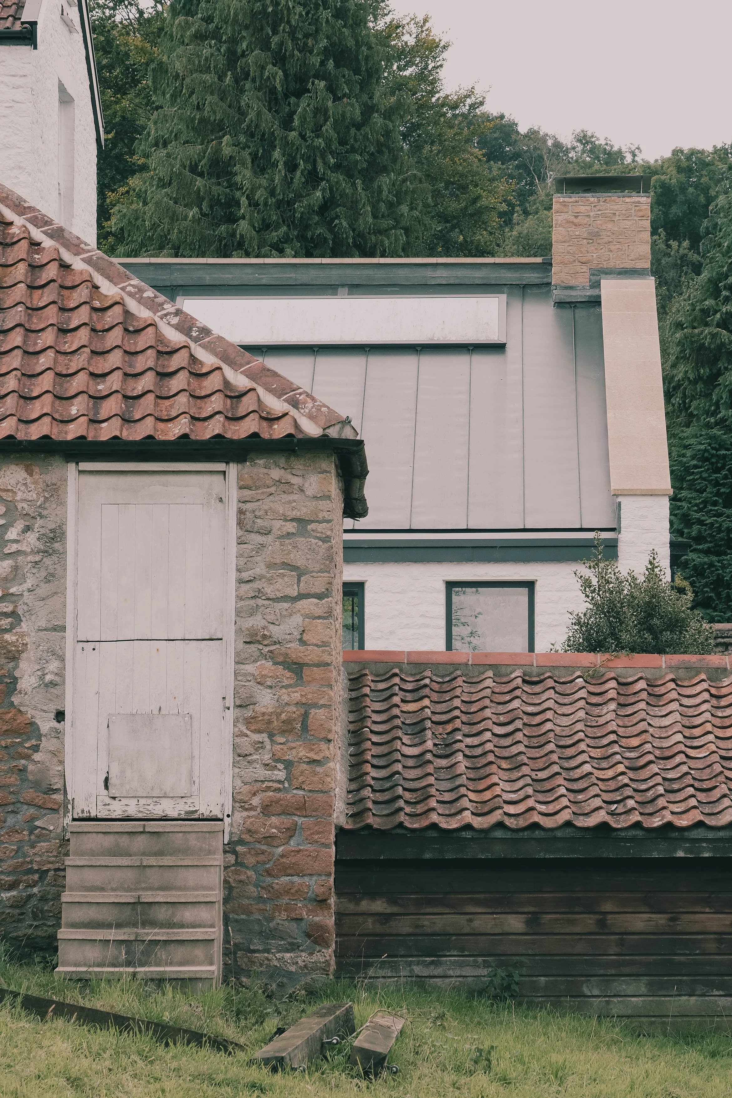 Layered scene showing an old stone outbuilding with a red‑tiled roof in front of a modern extension with a standing‑seam roof and chimney.jpg