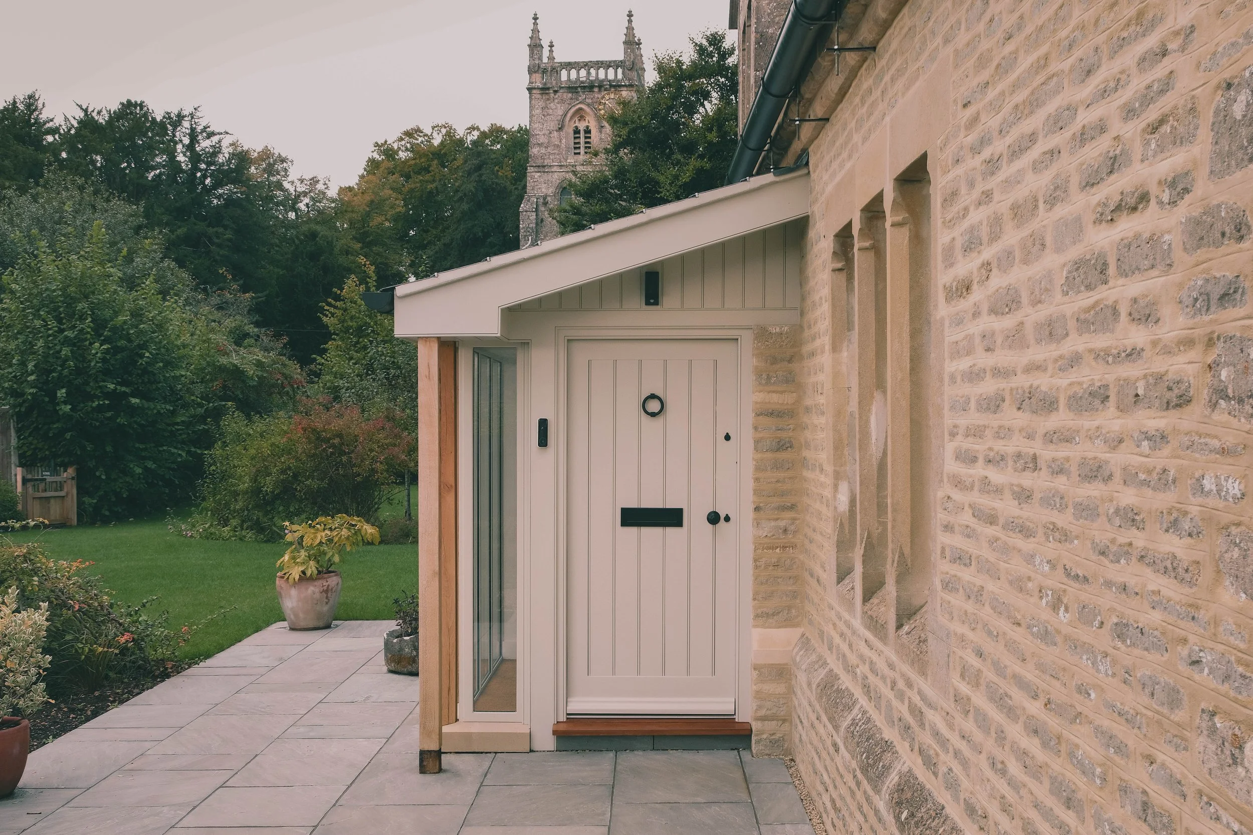 Stone house extension with a pale timber porch and panelled front door, set in a garden with a church tower visible beyond.jpg