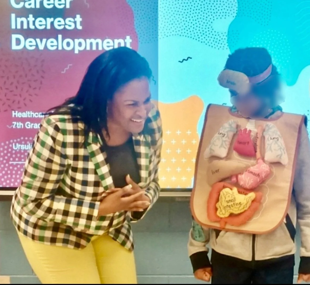 A woman and a child with a pretend torso model on a board in a classroom setting. The woman is laughing and the background features a colorful screen with text about career interest development.
