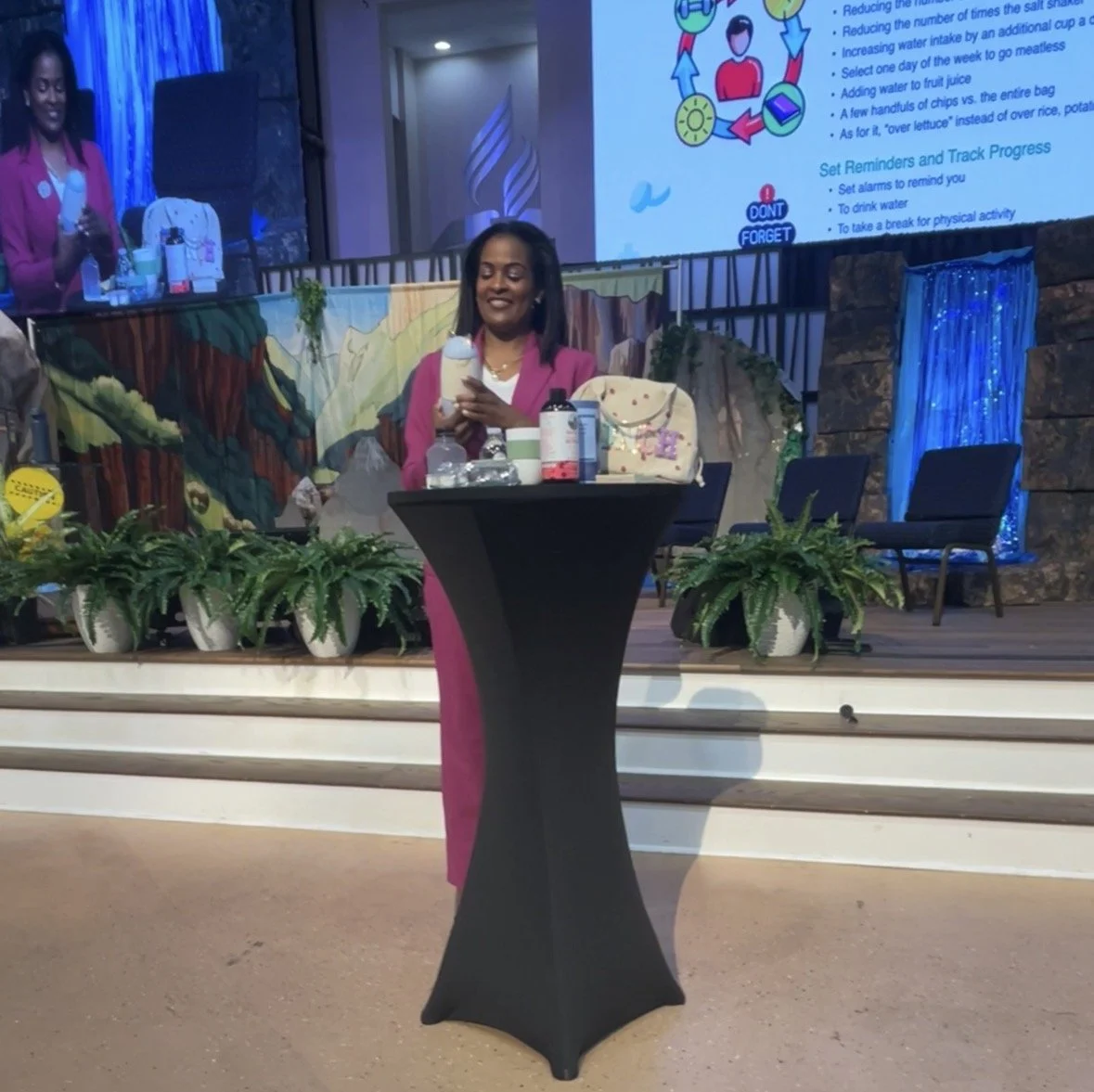 A smiling woman in a pink blazer and dress standing behind a tall black table on a stage, holding a container. The stage is decorated with potted plants, a painted backdrop, and chairs. A large screen behind her displays a presentation and a woman speaking on a smaller screen to the side.