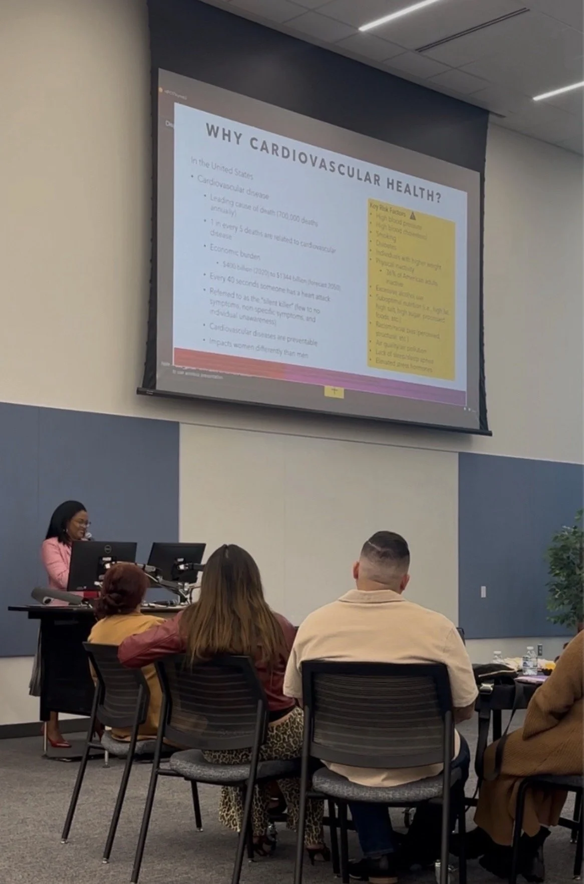 A classroom or conference room with a female instructor at a desk, presenting a slide titled 'Why Cardiovascular Health?'. Several students are seated at tables, facing the instructor and screen, listening attentively.