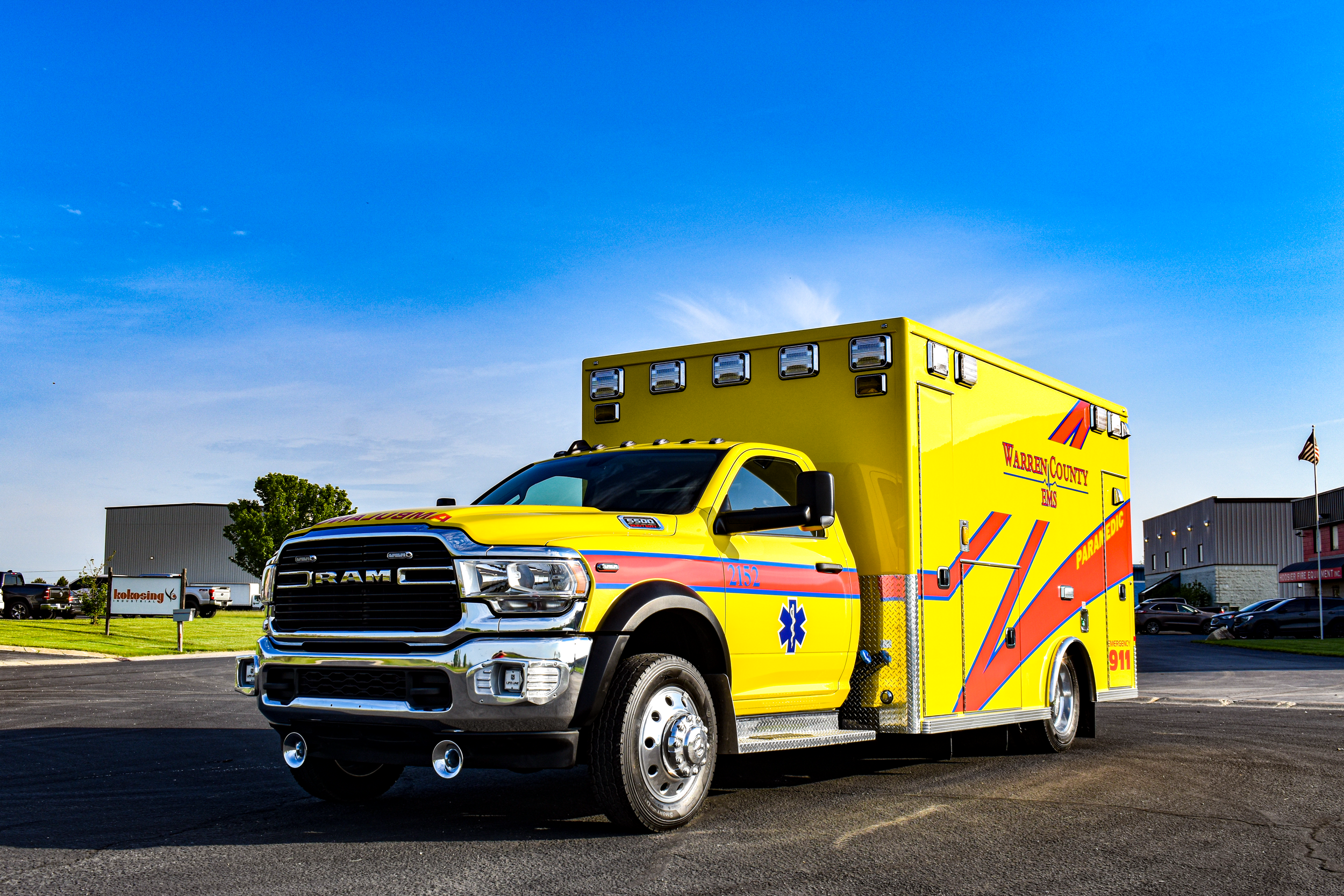 Yellow emergency ambulance vehicle with 'Warren County EMS' marking, parked in a lot on a clear day with bushes and buildings in the background.