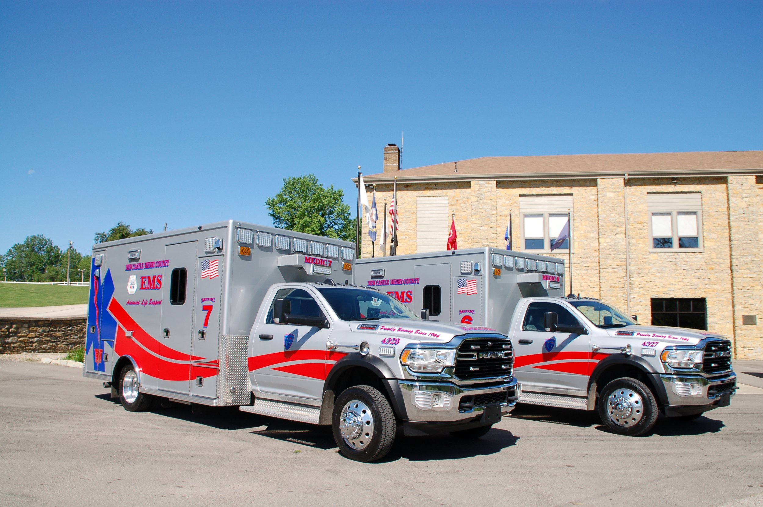 Two ambulance vehicles parked in front of a brick building with flags, clear blue sky, and some trees in the background.