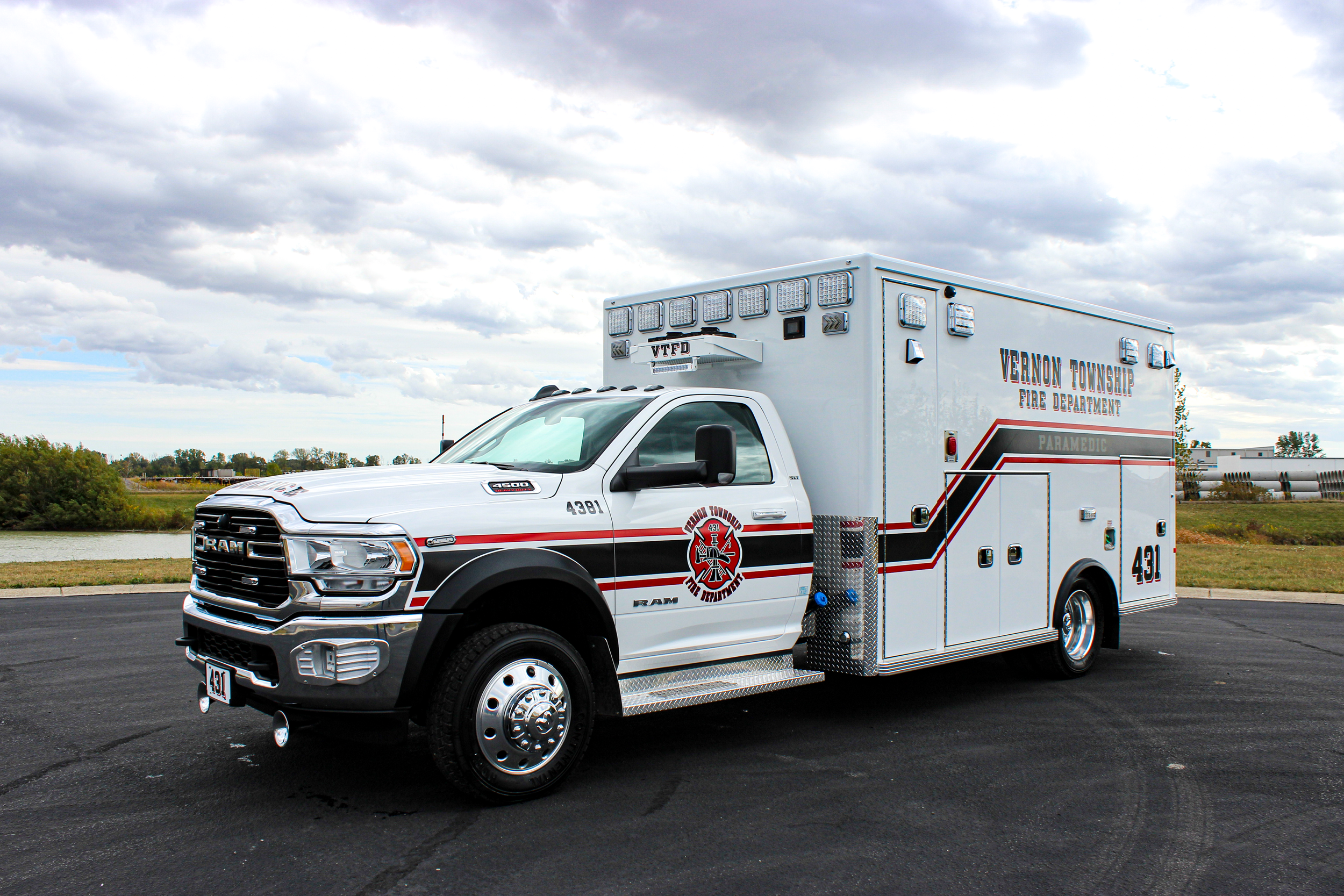 A white ambulance with red and black stripes and the words 'Vernon Township Fire Department Paramedic' on the side is parked on a paved surface near a body of water. The sky is cloudy and there are trees in the background.