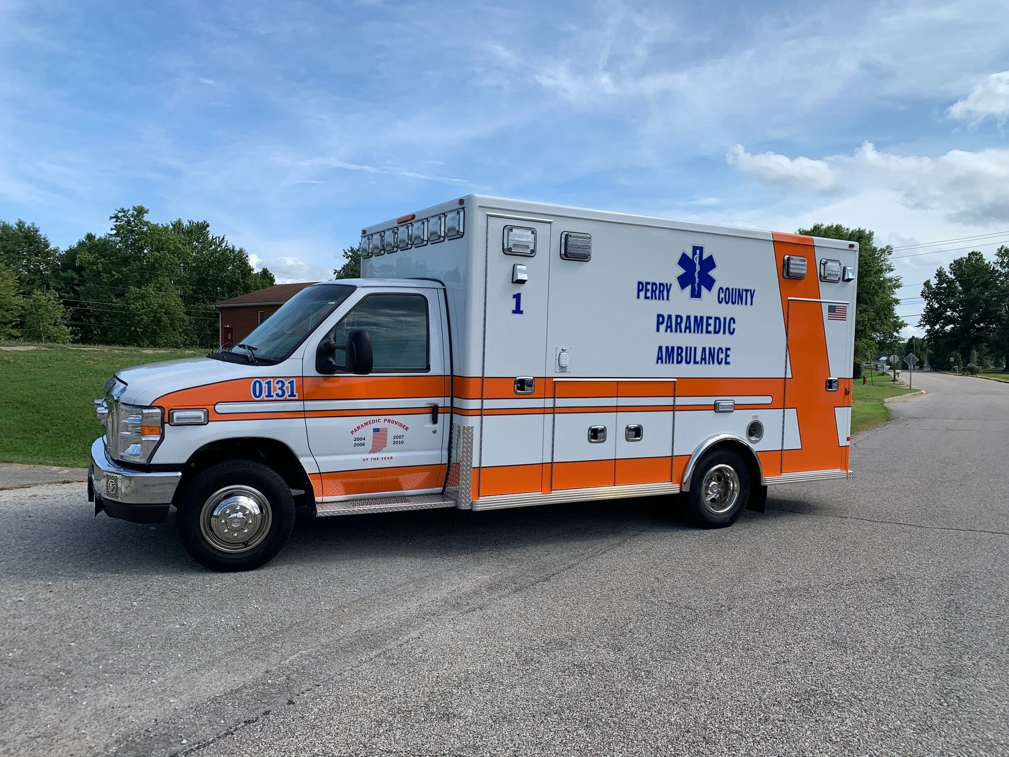 A Perry County paramedic ambulance parked on a paved street with trees and a house in the background under a partly cloudy sky.