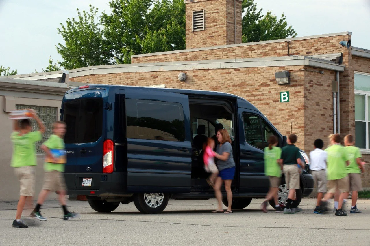 A group of elementary school children in uniform walking into a black van parked outside a brick school building, where a woman is helping a young girl enter the vehicle.