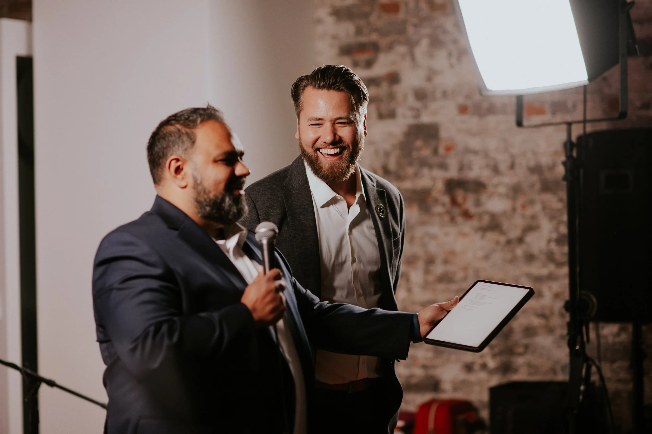 Raj Lulla and Ben Lueders - Two men dressed in business attire are conversing; one is holding a tablet, and they are smiling in a room with a brick wall, lighting equipment, and a microphone visible.