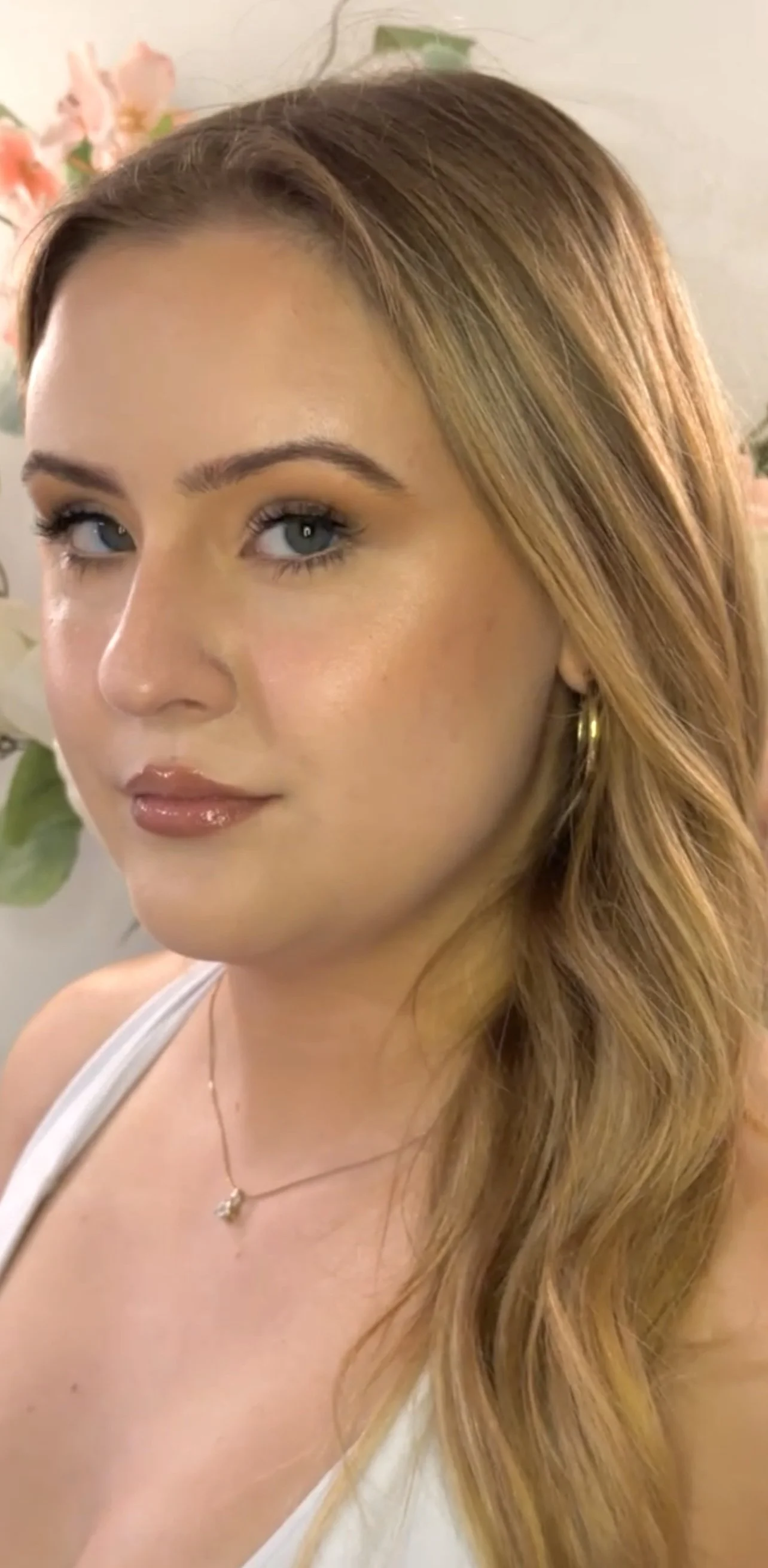 A young woman with light skin, blonde hair, and blue eyes, wearing a white top, a gold necklace, and gold hoop earrings. She is posing in front of a background with pink flowers and green leaves.
