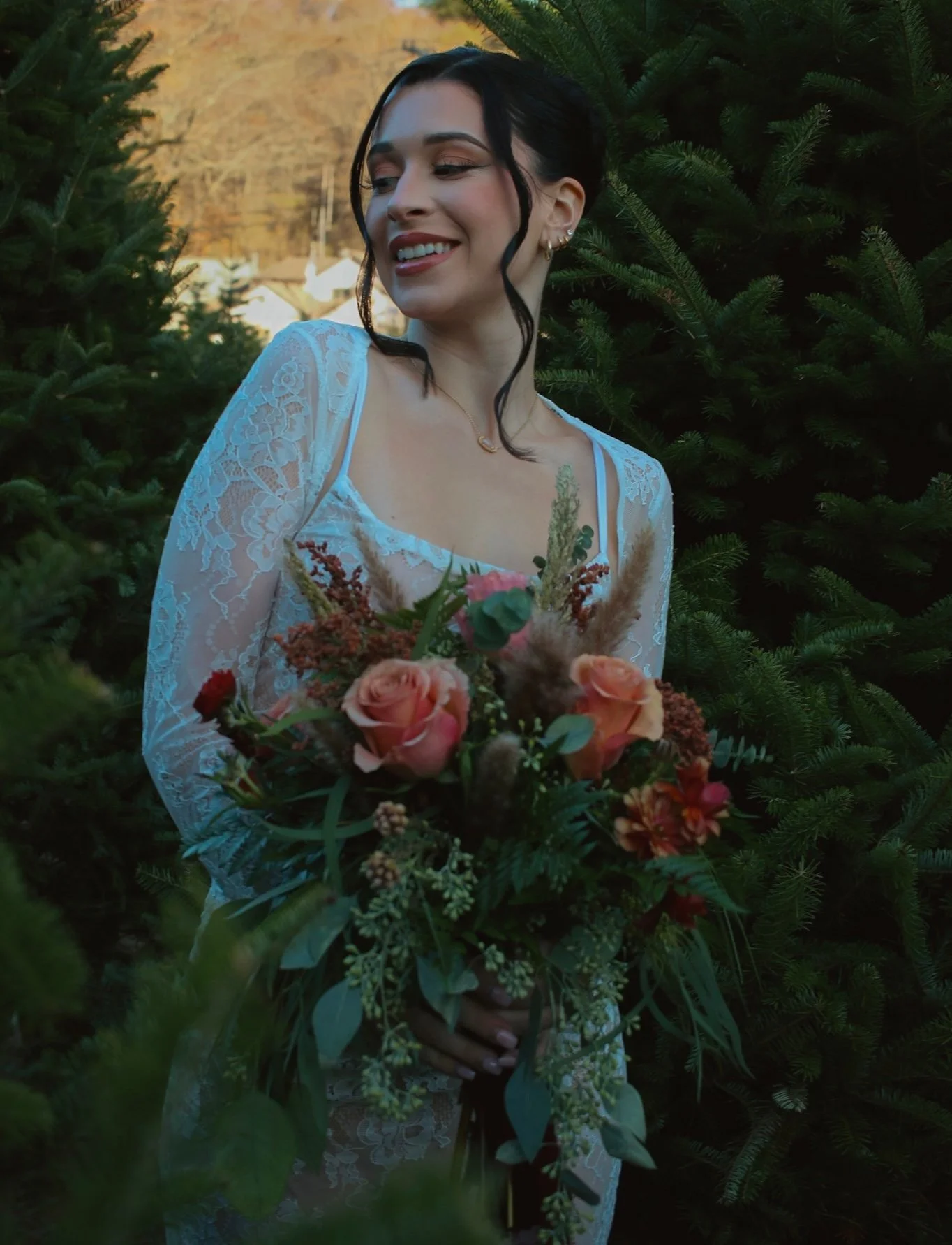 Woman in lace dress holding bouquet, surrounded by evergreen trees.