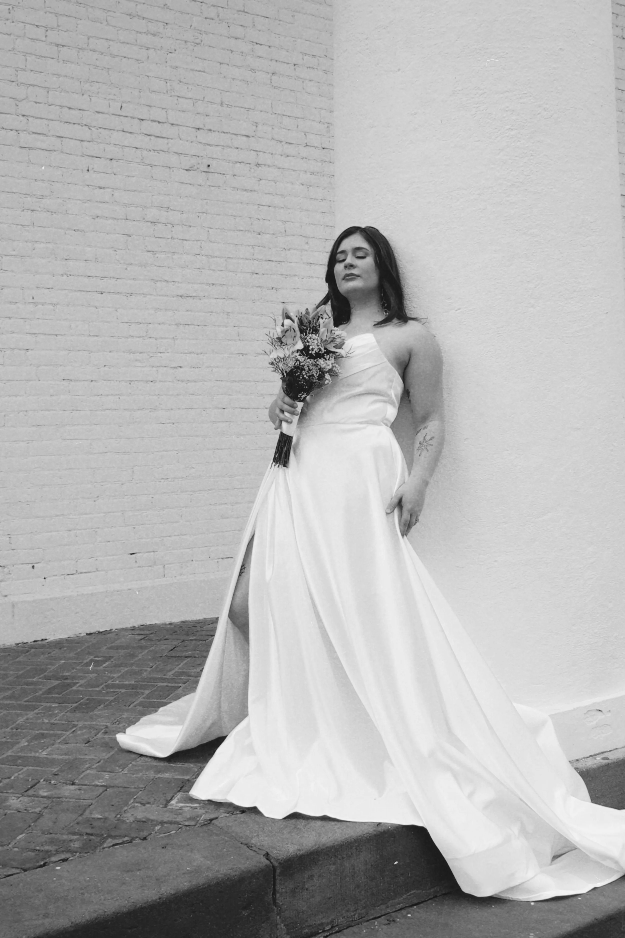 A woman in a wedding dress holding a bouquet of flowers, standing against a brick and painted wall in black and white.