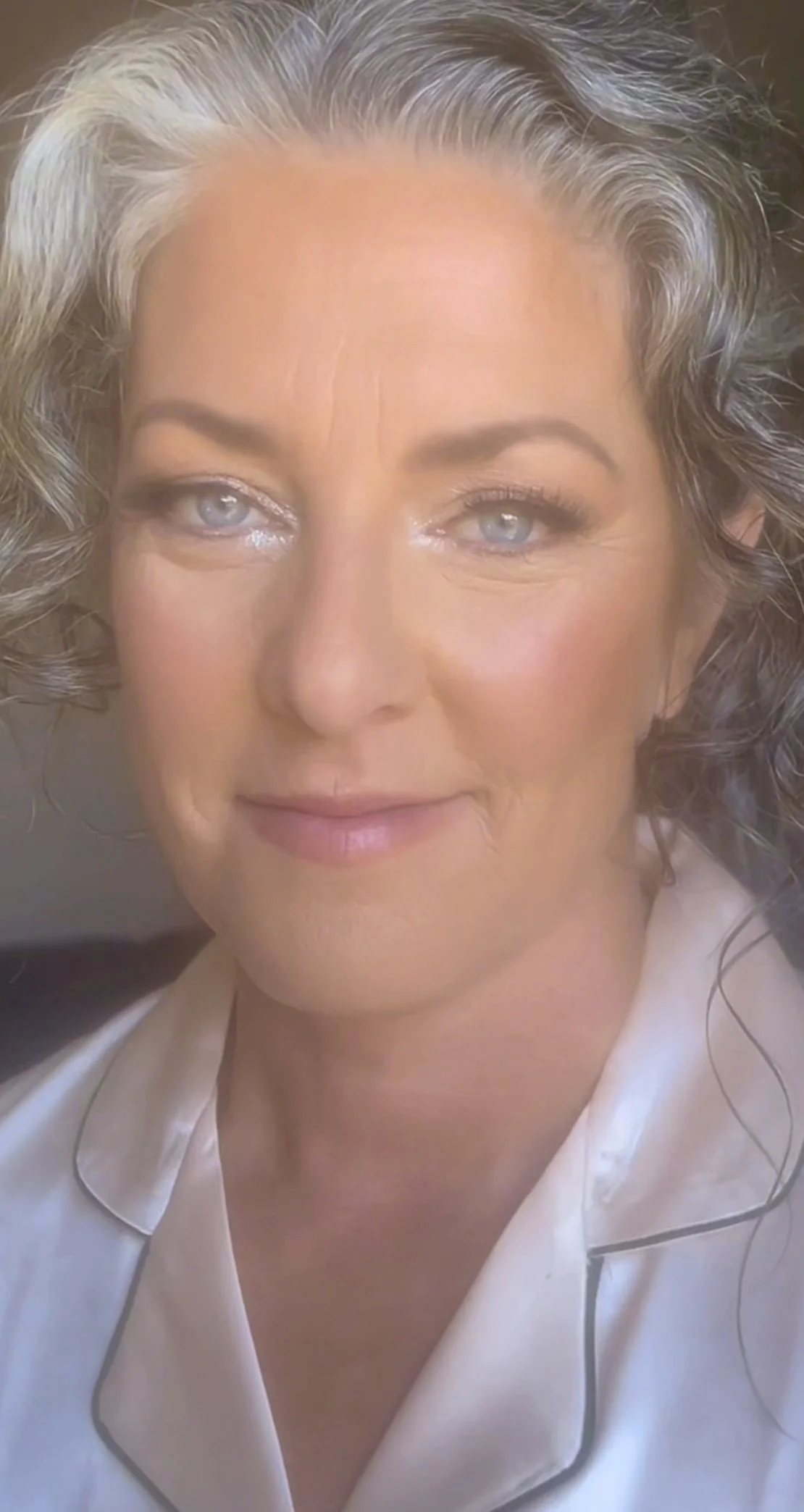 A close-up photo of a woman with light gray, curly hair and light eye makeup, wearing a white silk blouse with black piping.