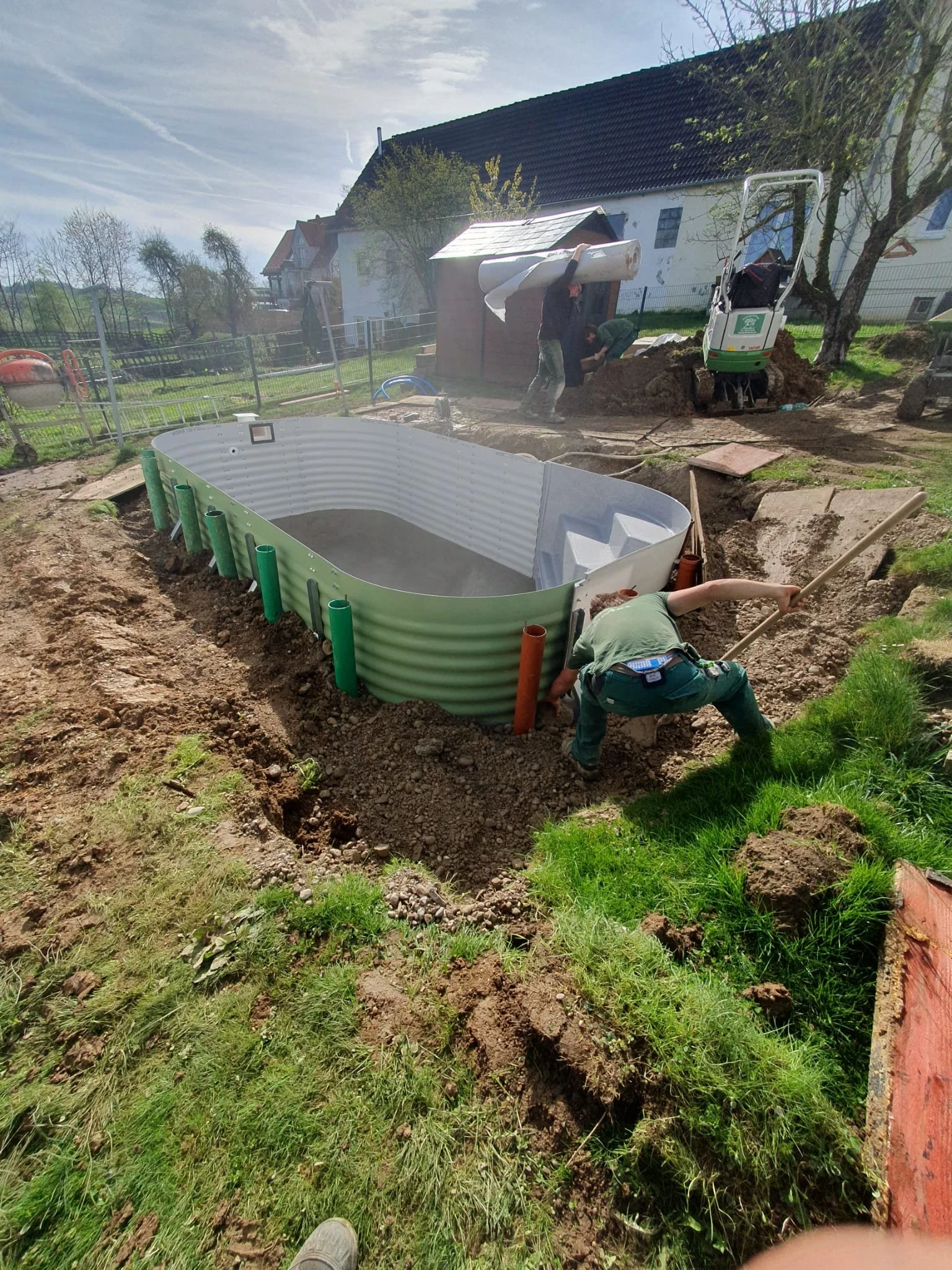 Ein Garten wird über einem Boden unterirdisch neu gestaltet, mit einem großen, ovalen witterungsbeständigen Pool, der gerade installiert wird. Mehrere Arbeiter sind an den Arbeiten beteiligt, einige beim Graben und andere beim Transportieren von Materialien.