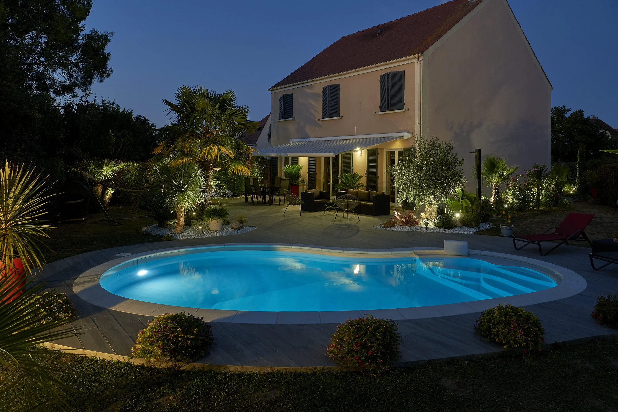 Ein beleuchteter Swimmingpool im Garten eines Hauses bei Nacht, umgeben von Büschen, Palmen und Gartenmöbeln.