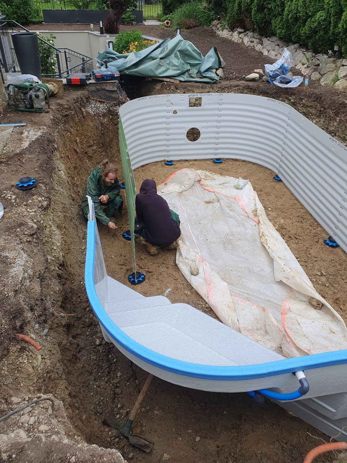 Zwei Personen bauen eine Pool-Fundament in einem Garten. Es sind vorgefertigte Poolwände vorhanden, die in einer Grube installiert werden.