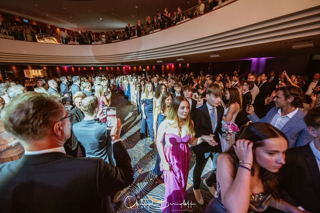 Festlicher Empfang mit einer großen Menschenmenge in einem eleganten Veranstaltungsraum, mit jungen und älteren Gästen in formeller Kleidung, einige nehmen Fotos auf.