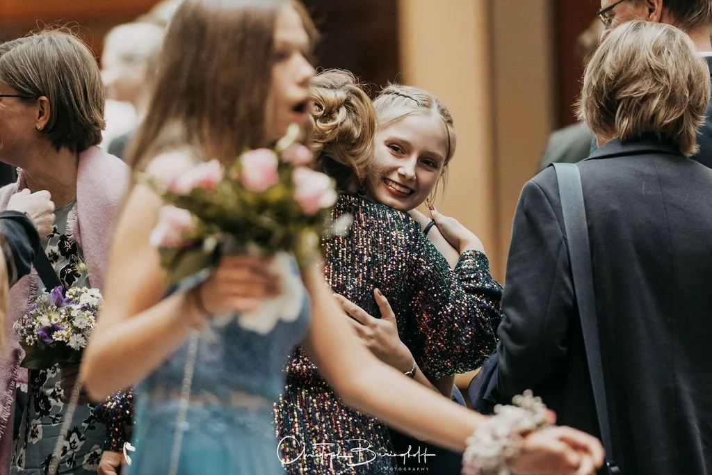 Junge Frau hält Blumenstrauß in der Hand, lächelnde junge Frau in Paillettenkleid umarmt eine andere Person auf einer Feier im Hintergrund.