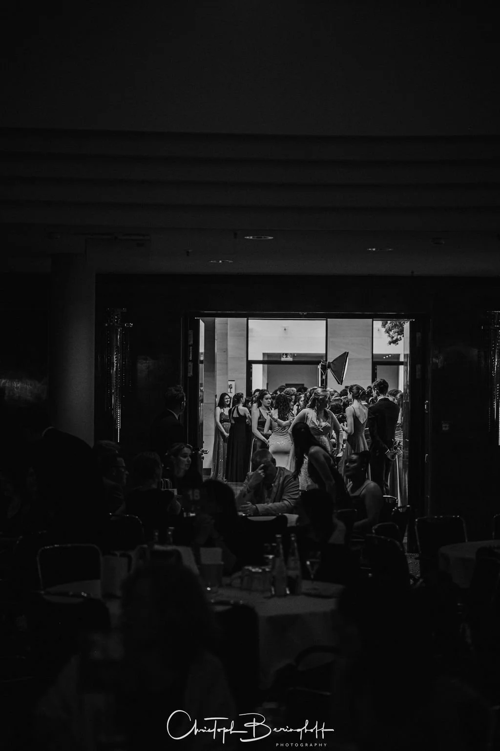 Schwarz-Weiß-Foto einer Veranstaltung mit Menschen, die im Hintergrund durch eine offene Tür ins Freie gehen. Im Vordergrund sitzen Menschen bei einem Empfang oder Dinner.