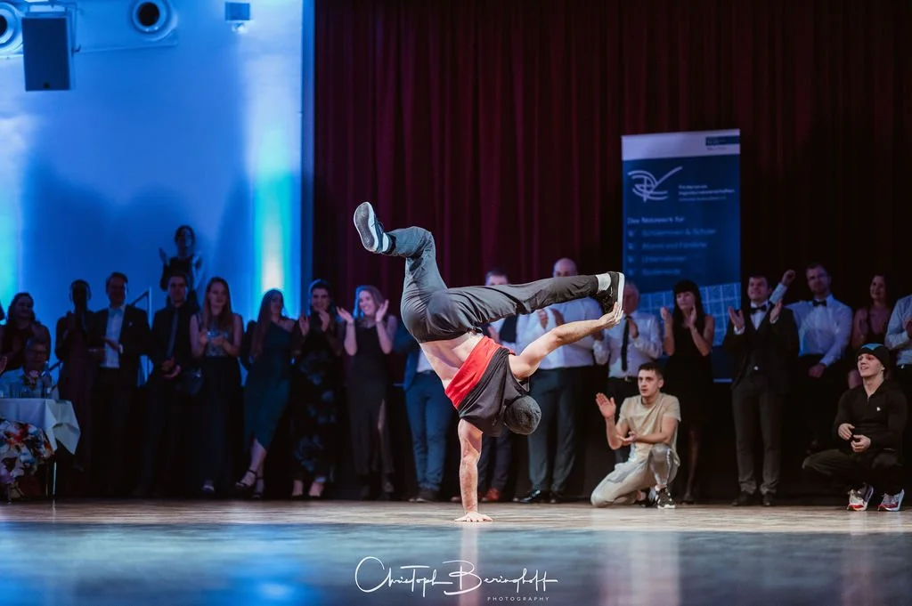 Breakdancer führt einen Handstand mit einem Bein in die Luft auf, umgeben von einem applaudierenden Publikum bei einer Veranstaltung.