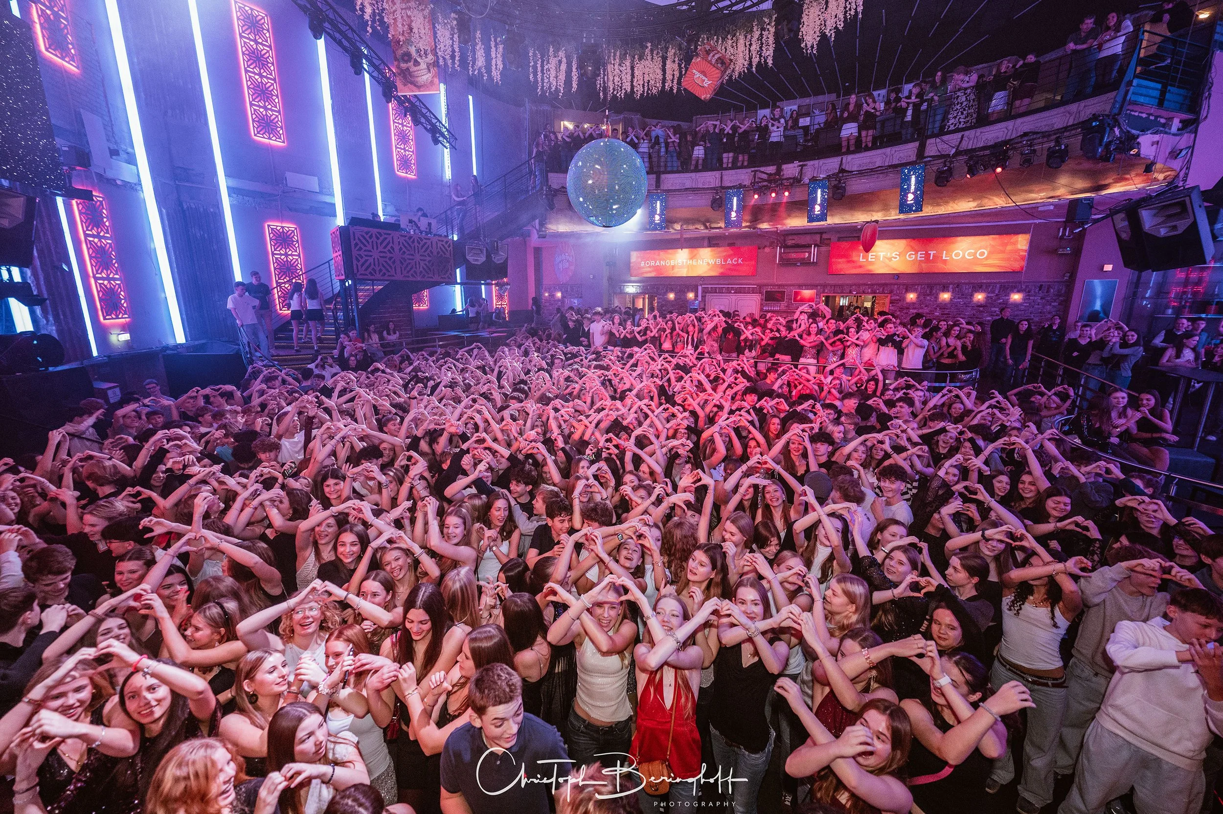 Menschenmenge in einem Club, die einen Herzschlag mit den Armen formen, auf einer Tanzfläche im dunklen, bunten Licht, mit Digitalanzeigen im Hintergrund