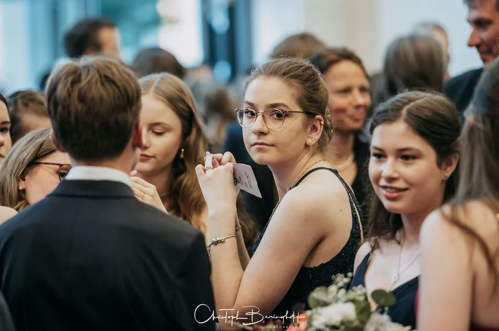 Menschenmenge bei einer formellen Feier, einige junge Frauen und Männer, die sich unterhalten, mit einem jungen Mädchen in der Mitte, das an der Kamera vorbeischaut.