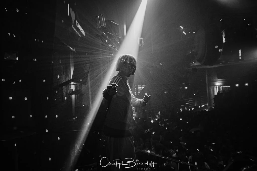 A person stands on a stage in a dark room, illuminated by a single bright beam of light, with sound and lighting effects in the background, while an audience watches.