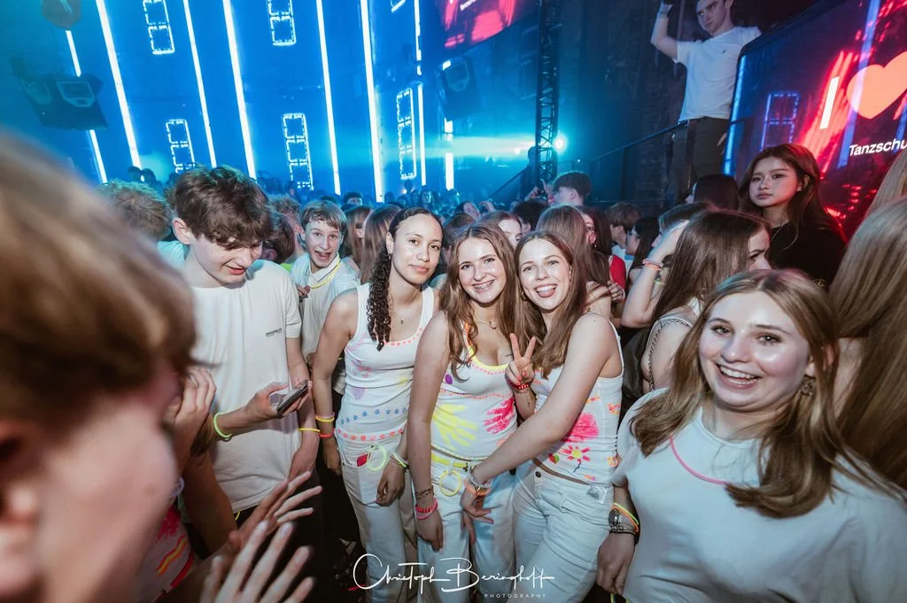 Young people are partying at an event with a neon-lit stage and LED lights; everyone is wearing white T-shirts or white clothing.