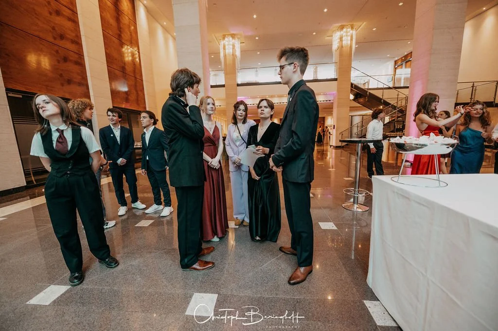 Eine Gruppe junger Menschen in formeller Kleidung, die sich unterhalten, in einer eleganten Hotelhalle oder Veranstaltungsraum, während in der Hintergrund andere Gäste und ein Buffet zu sehen sind.