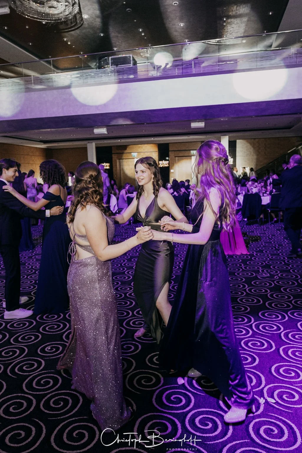 Junge Frauen tanzen auf einem festlichen Ball im eleganten Abendkleid unter bunten Lichtern, umgeben von weiteren Gästen.