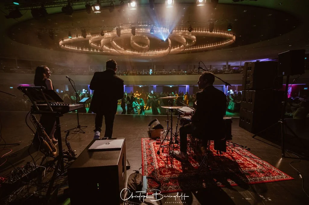 Musiker auf einer Bühne in einem dunklen Veranstaltungsraum mit Blick auf das Publikum, das bei einer Live-Performance tanzt, mit farbigem Licht- und Stage-Design.