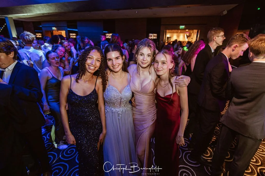 Eine Gruppe von vier jungen Frauen in eleganten Kleidern posiert auf einer Party, umgeben von einer Vielzahl anderer feiernder Gäste in einem festlich beleuchteten Raum.