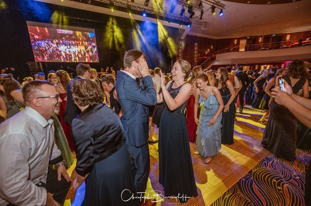 Menschen auf einer Tanzfläche bei einer Party, lachen und feiern in einem festlich beleuchteten Veranstaltungsraum.