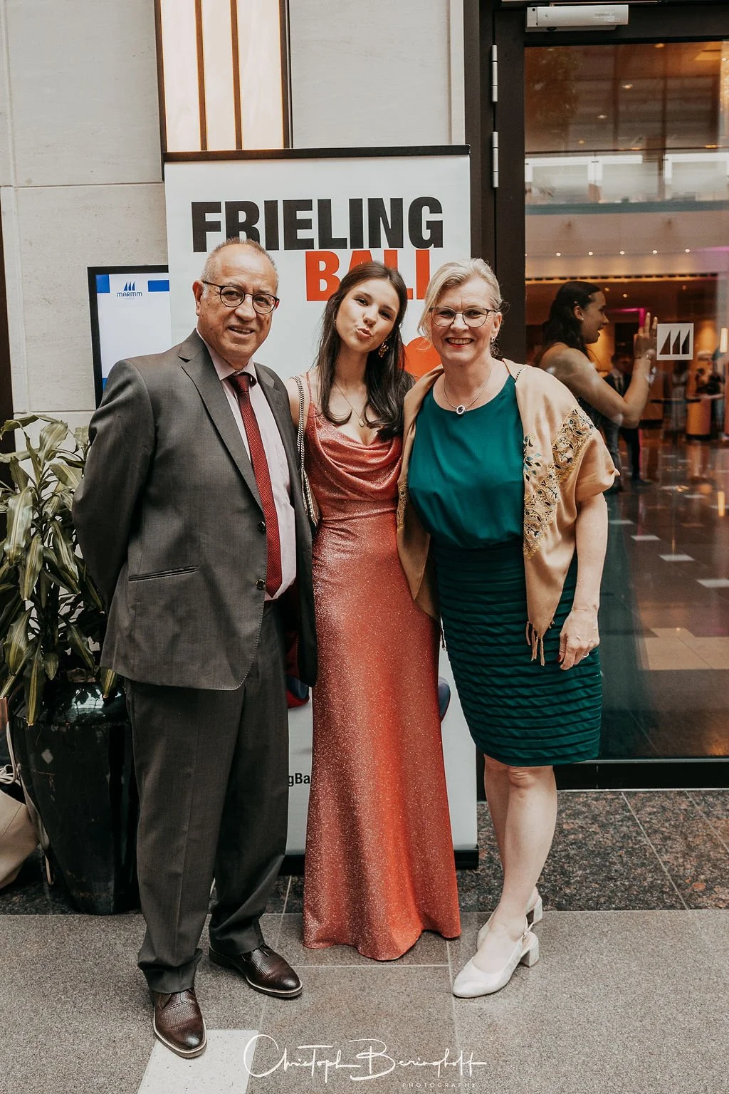 Drei Menschen im formellen Gesprächsfest bei einer Veranstaltung, stehend vor einem Poster mit der Aufschrift 'FRIELING BALL', lachend und in elegante Kleidung gekleidet.