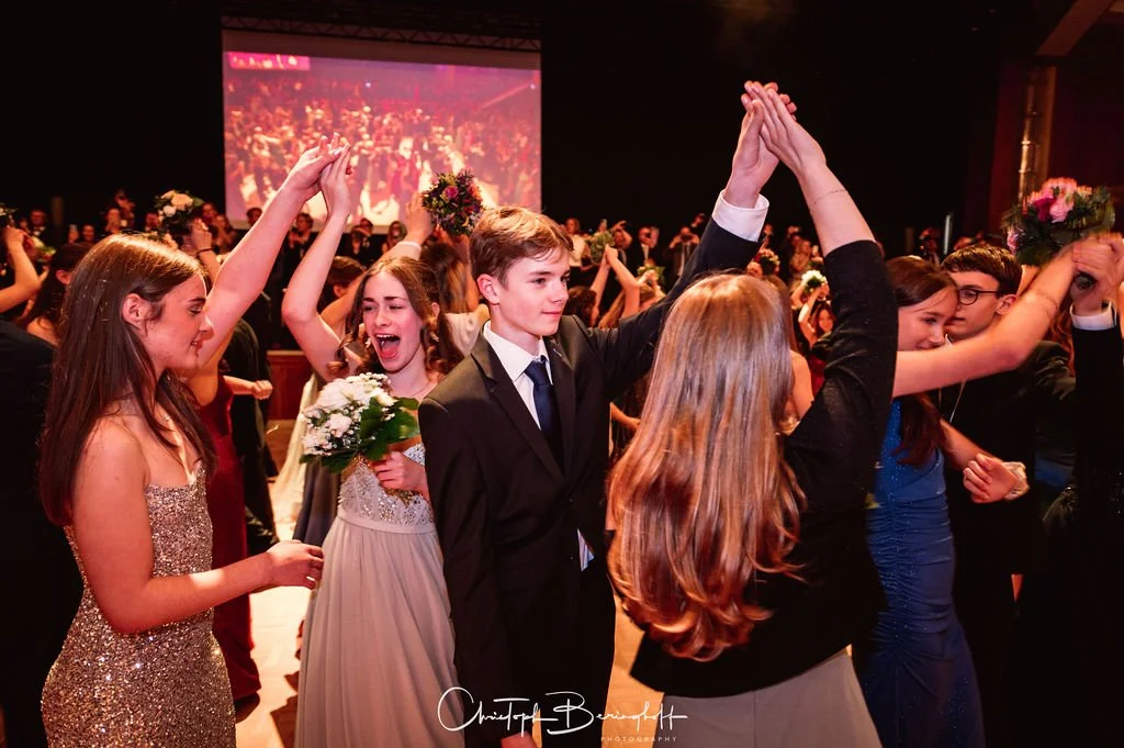 Jugendliche tanzen auf einem festlichen Ball, einige halten Blumen und feiern miteinander.