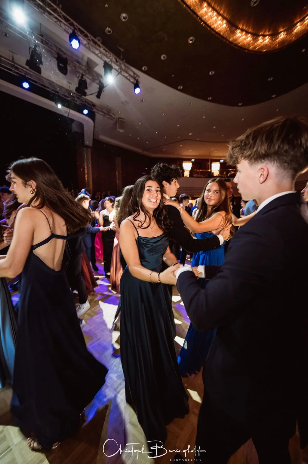 Jugendliche tanzen auf einer Party in eleganten Kleidern und Anzügen in einer großen, gut beleuchteten Halle.
