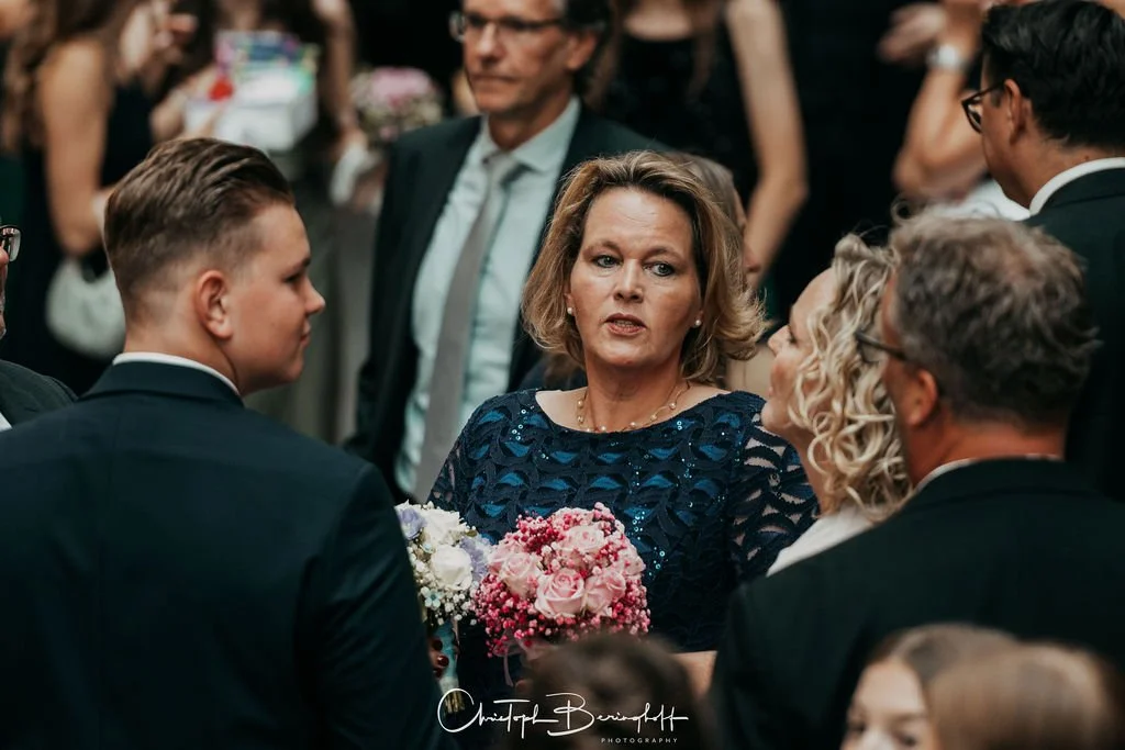 Ältere Frau im blauen Kleid spricht mit jüngeren Personen auf einer formellen Veranstaltung, während sie eine Blumenstrauß hält.