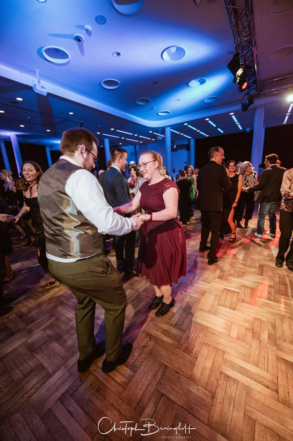 Menschen tanzen auf einem festlich beleuchteten Tanzparkett, feiern und lachen in einem modernen Veranstaltungsraum.