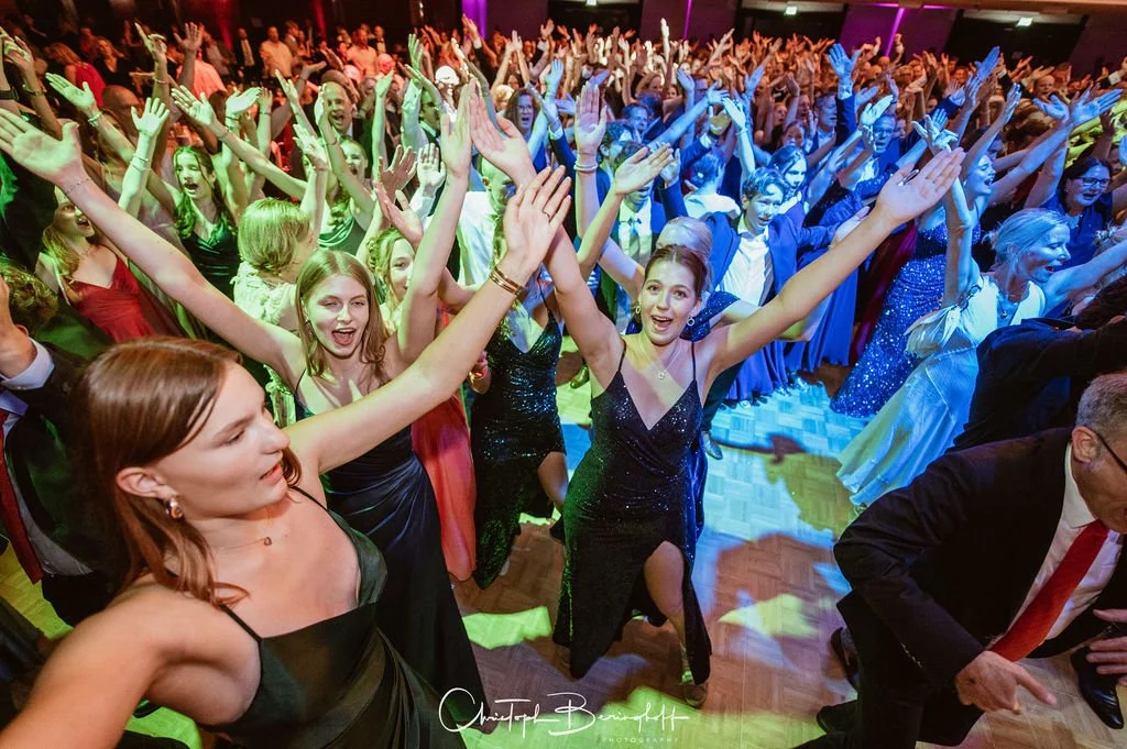 Menschen feiern auf einer Tanzparty, alle haben die Arme in die Luft, bunte Partybeleuchtung, festliche Kleidung.
