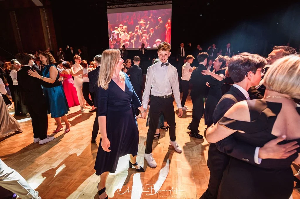 Tanzende Menschen bei einer Gala oder Ball, ältere Damen in eleganten Kleidern, junge Männer in Anzügen, große Projektionsleinwand hinter der Tanzfläche, festliche Atmosphäre.