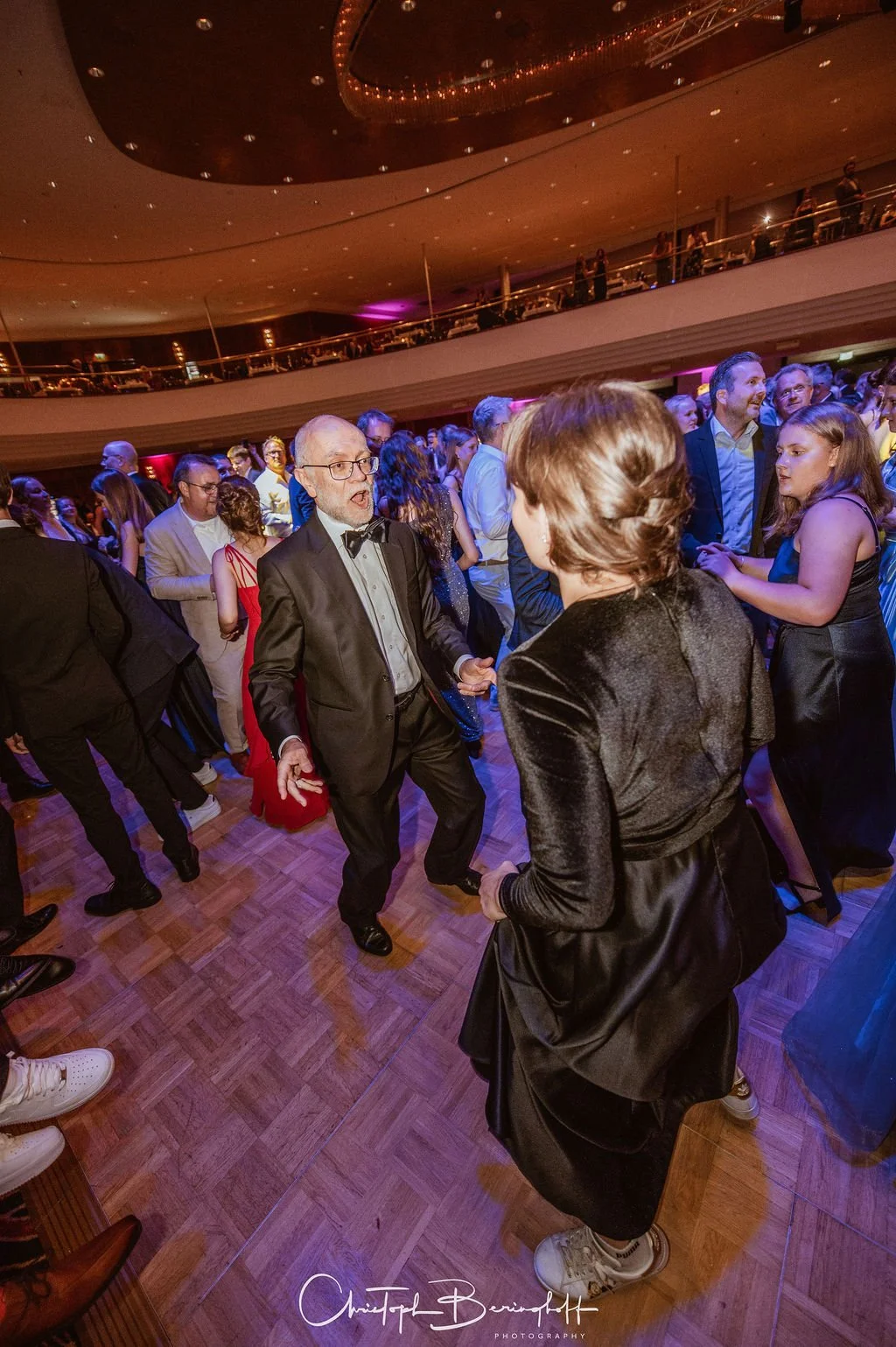 Menschen tanzen bei einer Feier in einem großen Saal mit mehreren Etagen und warmem Licht.